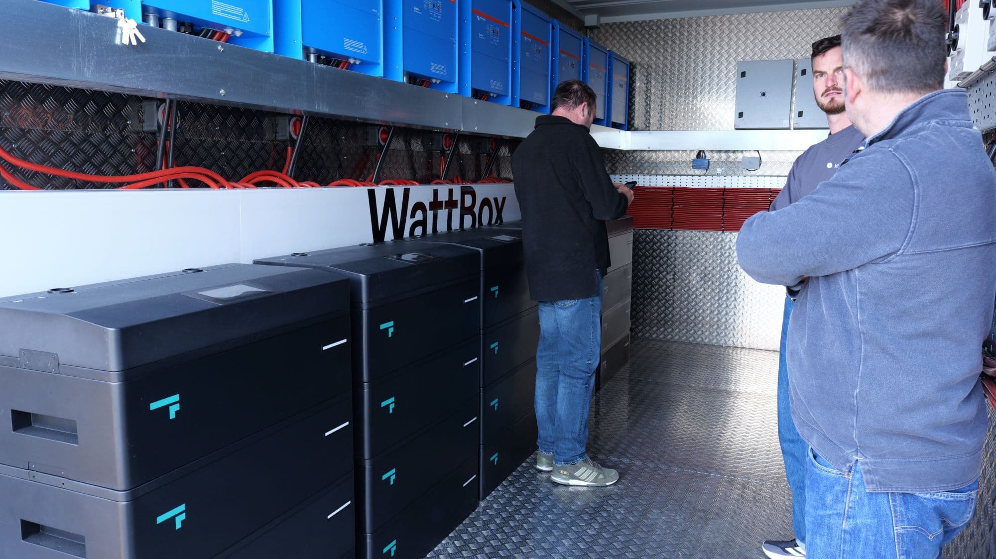 Engineers inside a WattBox container, beside the battery stacks and a row of Victron inverters